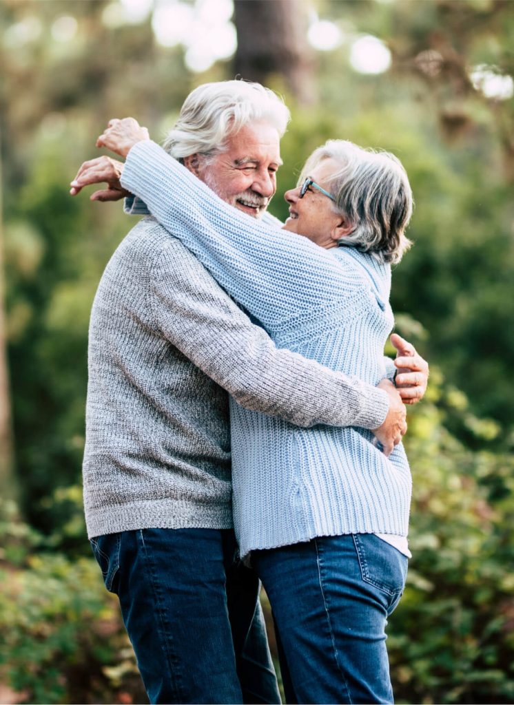Two older adults with gray hair, wearing sweaters and jeans, embrace and smile at each other outdoors in a green, natural setting.