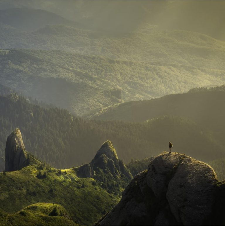 A person stands on a rocky outcrop overlooking a vast, sunlit landscape of green hills and distant forests under a hazy sky.